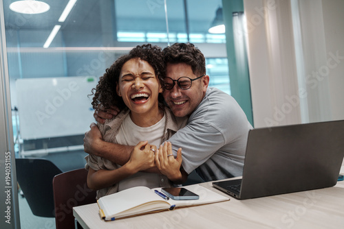 Obraz w ramie Happy Colleagues Share Warm Hug. Bright Office Scene Showcasing Genuine Emotion And Team Closeness