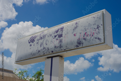 Foto A faded outdoor billboard sign mounted on a pole features a blank, weathered advertising panel with peeling paint and ghosted graphics against a bright blue sky with scattered clouds