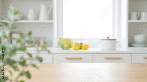 Bright and airy modern kitchen interior with blurred foreground plant, featuring white cabinets, light wood countertop, and fresh produce by a sunlit window