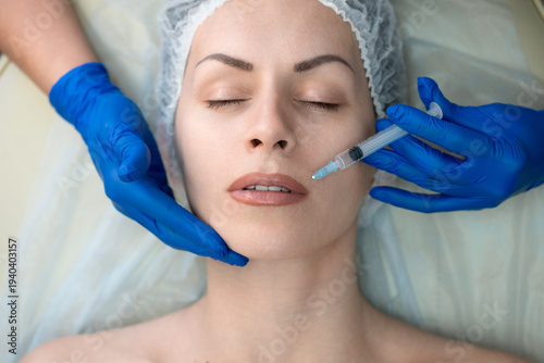 Female head in medical cap lying on a couch and get professional cosmetic injection in nasolabial fold area, aesthetic medicine procedure with doctor hands in blue gloves, close-up