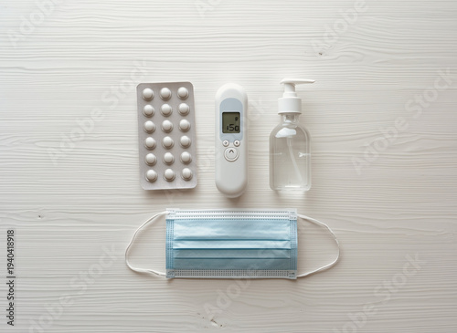 High-angle flat lay shot of essential healthcare and wellness items: generic medical pills in blister packs, a digital thermometer, a small bottle of hand sanitizer, and a generic blue face mask on a 