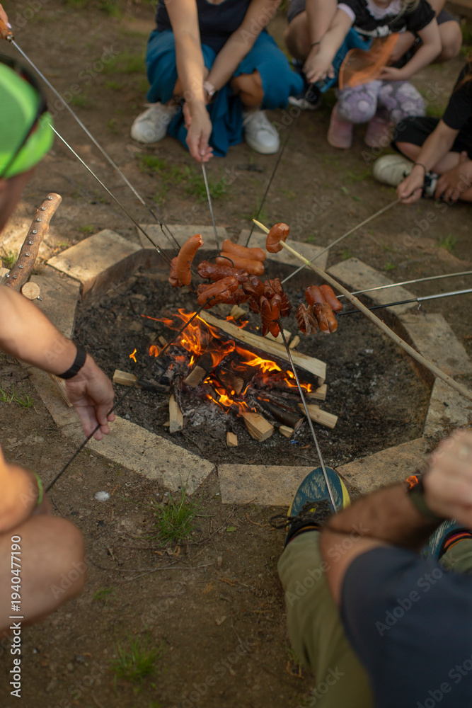 Obraz premium Group grilling sausages around firepit at twilight, parents and kids tending skewers, orange flame flicker, picnic style seating. People celebrate the traditional Czech holiday Carodejnice outdoors.