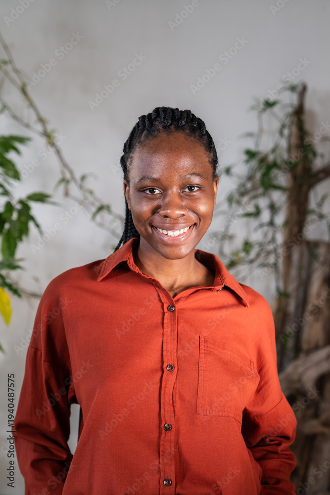 Fototapeta premium Positive confident African American woman posing smiling at camera with optimistic calm expression. Black female surrounded by green houseplants and natural decorations poses at home garden space.