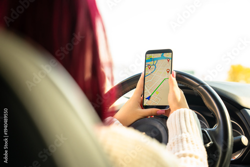 Young woman sitting inside a car, studying navigation on her smartphone with a concerned expression after getting lost in an unfamiliar city.
