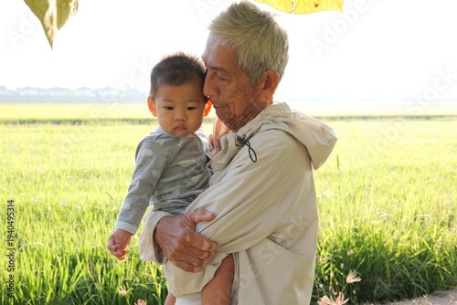 An elderly Asian grandfather holding his baby grandson outdoors in a green rice field during warm natural sunlight. 