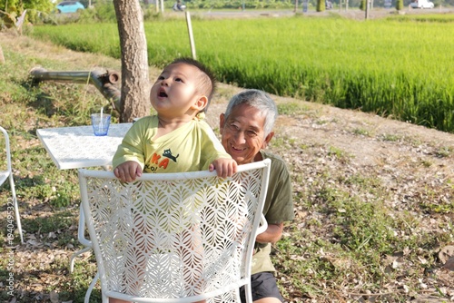 A smiling Asian grandfather playing with his baby grandson outdoors near a green rice field. 