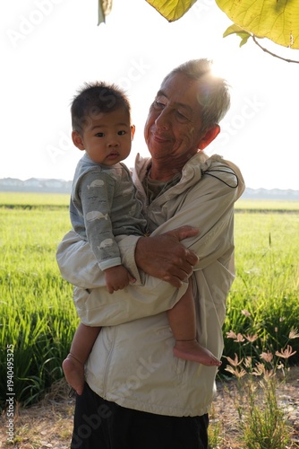 An elderly Asian grandfather gently holding his baby grandson outdoors in a green rice field during warm sunset light. 