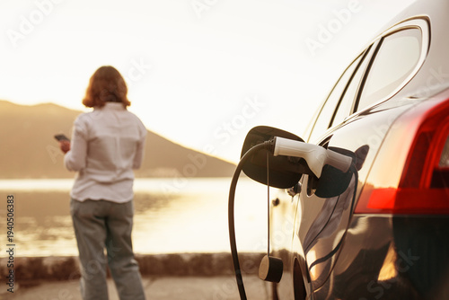 Photos Woman with smartphone next to charging electric car on the seacost during summer