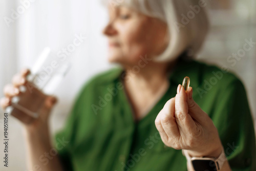 Senior woman holding pill and glass of water while sitting at home. Mature caucasian female taking medication, illustrating healthcare, treatment and illness management in retirement lifestyle