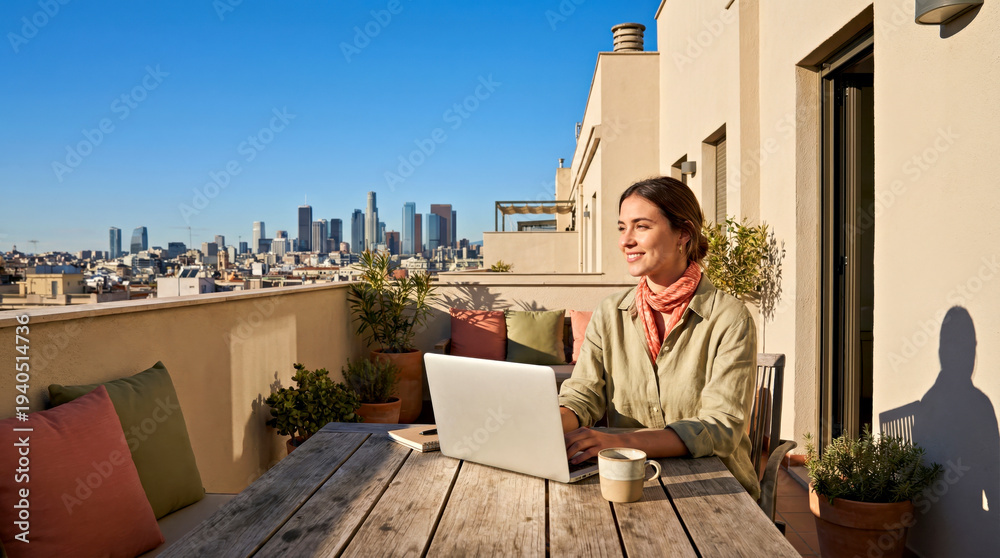 Obraz premium Woman working on laptop at outdoor table on balcony with city skyline view, potted plants and cushions visible in bright sunlight