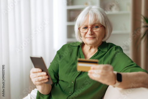 Senior woman sitting on sofa at home using smartphone and holding credit card. Mature caucasian female making online payment or shopping on mobile device during retirement lifestyle