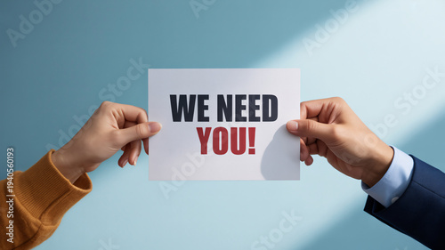 Two hands holding a white card with 'WE NEED YOU!' text, symbolizing a call for recruitment or participation.