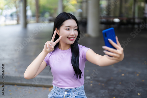 фотографія Young Asian woman smiles taking selfie with smartphone making peace sign gesture