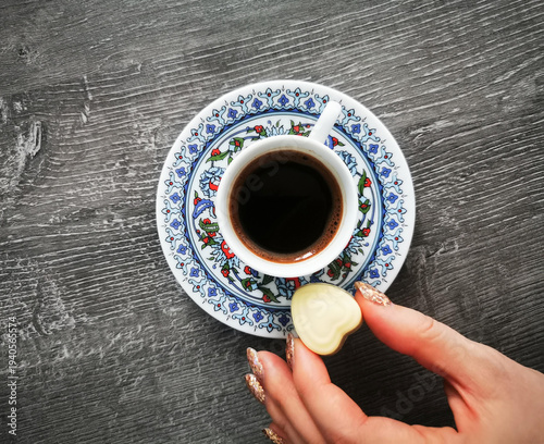 Photos A decorative cup of coffee sits on a patterned saucer as a hand places a chocolate heart beside it