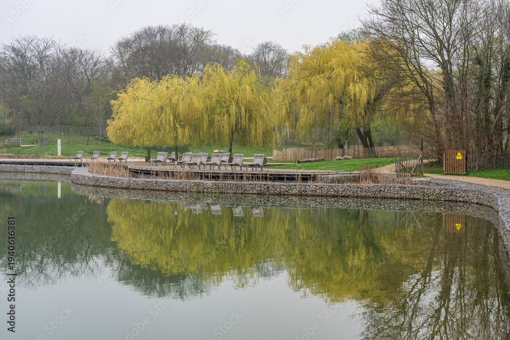 Obraz premium Gennevilliers, France - 03 08 2026: Chanteraines park. Nature in spring season. Reflection of weeping willow trees, wooden chairs and vegetation with cloudy sky on the lake