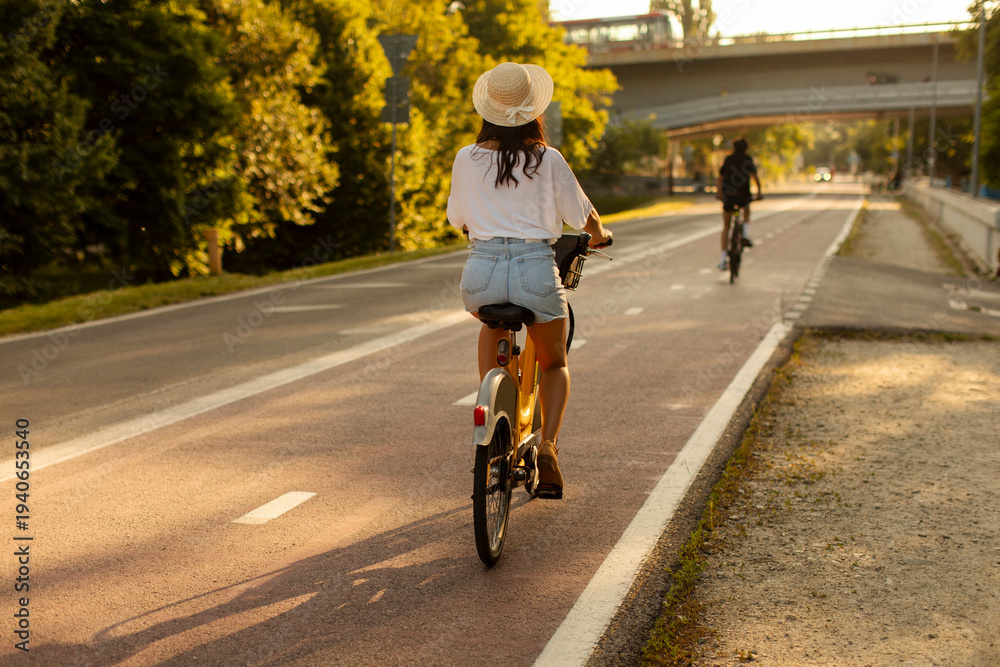 Fototapeta premium Back view of woman riding bike on path with trees lining the way, lady cycling on sunny day, enjoying fresh air and nature
