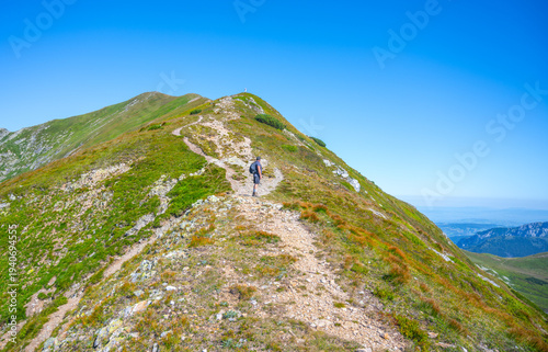Hikers traverse the winding path on Bystra Mountain, enjoying clear blue skies and the lush greenery typical of the Tatras in Slovakia. A serene adventure through nature's beauty awaits.
