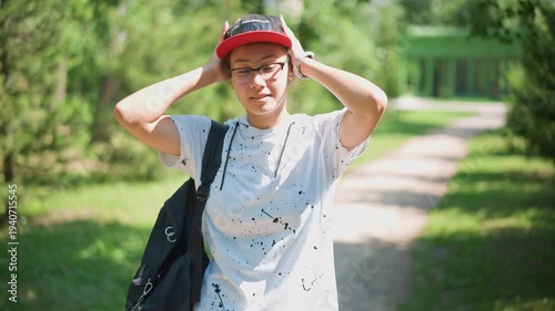 portrait person adjusting cap on path playful grin and glasses catching light, creator and influencer vibes, confident pose with casual hoodie and backpack, green park backdrop, summer warmth