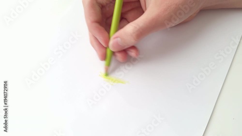 A young man using a light green colored pencil to draw a soft stroke on a clean sheet of white paper. Close up.