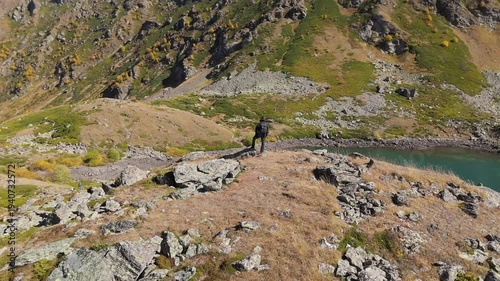 Lone figure stands atop rugged terrain beside a turquoise lake. The person gazes out at the vast, sunlit valley beyond. Sunlight glints off the water and rocks in golden hues