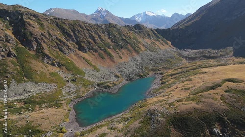 Aerial view reveals a turquoise lake nestled in rugged mountains. Snow-capped peaks frame the serene, glacial-fed waters. Golden grasses surround the valley, hinting at autumn's approach