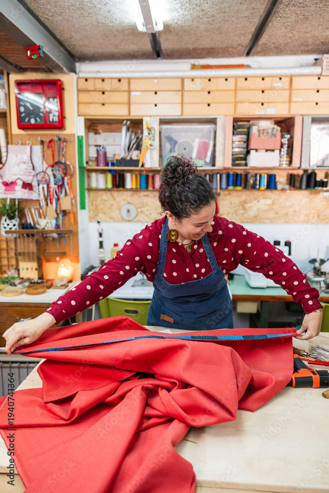 Naklejka premium Woman artisan measuring red fabric with a tape measure on a wooden table, preparing it for a custom upholstery project. Celebrating craftsmanship and small business