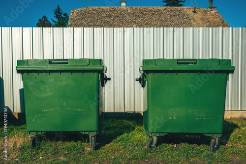 Two large green wheeled garbage containers (dumpsters) side by side on grass, lids slightly ajar, positioned in front of a tall silver corrugated metal fence