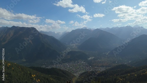 Highland vista frames a valley town nestled below. Distant peaks rise above layered mountain ranges. Blue sky with wispy clouds bathes the scene in light