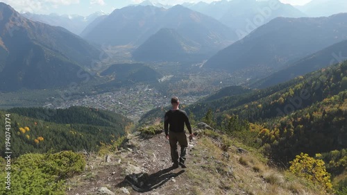 Two hikers pause atop a rocky mountain peak. One sits, resting with a backpack beside them. The other stands, gazing at distant valleys and peaks. Clear blue sky with wispy clouds above