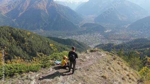 Hiker pauses on mountain ridge, overlooking vast valley. Another hiker sits nearby, enjoying the view. Sunlit peaks stretch behind them, framed by autumn trees