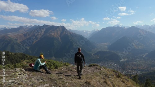 Two hikers pause atop a rocky mountain peak. One sits, resting with a backpack beside them. The other stands, gazing at distant valleys and peaks. Clear blue sky with wispy clouds above