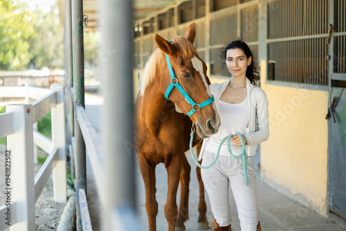 Fotografía Smiling Armenian female horse farm worker takes horse out of stall, holds animal by bridle and leads pet for walk