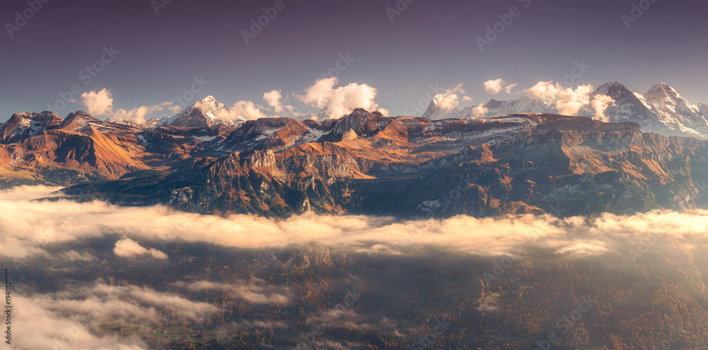 Fototapeta premium Panoramic aerial view of the Swiss Alps with snow-capped peaks and low clouds at sunset. Bright evening light and clear blue sky over rugged alpine mountains in Switzerland. Top view. Landscape