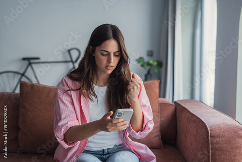 A frustrated young woman sits on her couch at home, looking angrily at her smartphone screen, feeling unhappy and dissatisfied after receiving bad news or dealing with a broken device