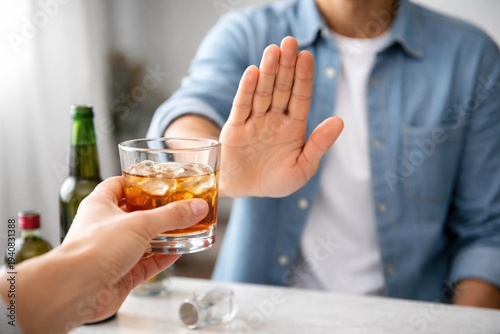 refusing alcohol and sobriety decision, Young caucasian man raises hand to refuse glass of alcohol offered by another person. Whiskey and bottles on table. Concept of sobriety, alcohol refusal and hea