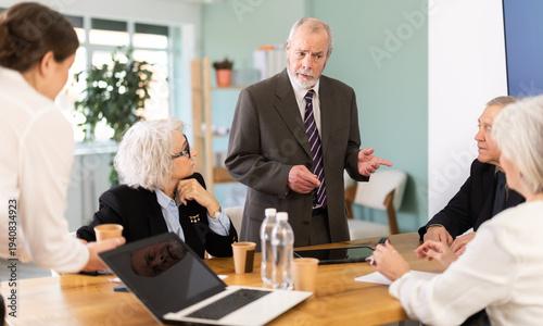 Obraz na płótnie Irritated elderly male boss reprimanding upset subordinates sitting at table in