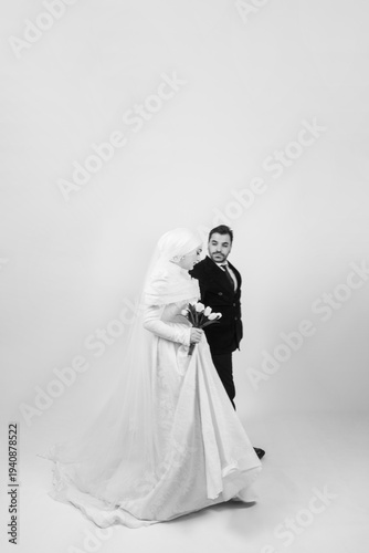 Minimal black and white wedding portrait of a Muslim bride and groom walking together in a studio. The bride wears a modest lace wedding dress with hijab and holds a bouquet, while the groom walks 