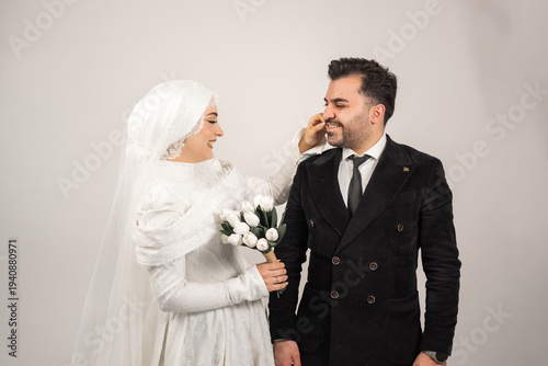 Romantic moment between bride and groom in a studio wedding portrait. The bride wearing a modest white wedding dress and veil gently touches the groom’s face while holding a bouquet of white tulips.