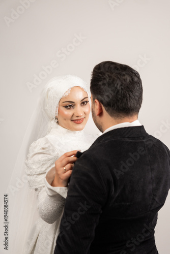 Beautiful bride gently adjusting the groom’s suit collar in an intimate wedding moment. The bride wears a modest lace wedding dress with a delicate veil and elegant makeup while smiling warmly 
