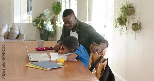 African American father guiding school-age son prompted by homework on laptop writing at home table