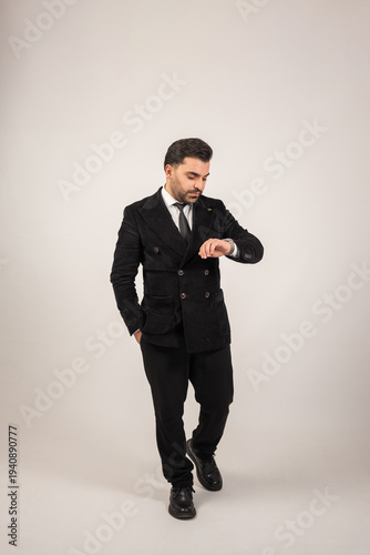 Full body portrait of a fashionable businessman checking his wristwatch while standing in a clean studio setting. The minimalist background emphasizes his modern style and professional look.