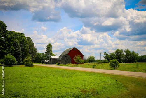 A red barn stands near a quiet road surrounded by lush green grass and trees under a wide blue sky filled with fluffy white clouds in a peaceful rural landscape