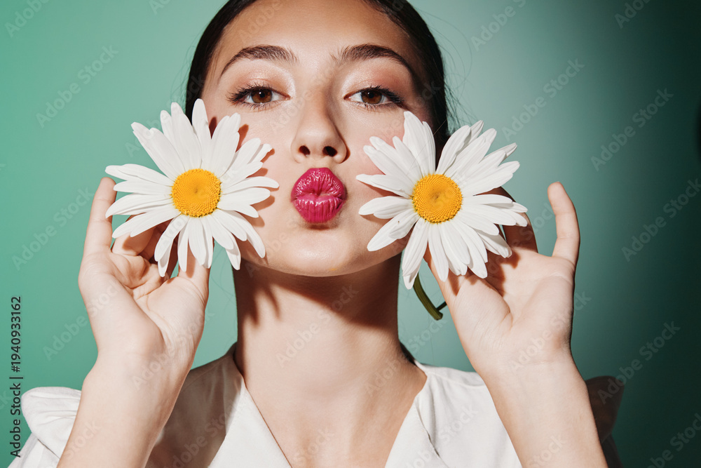 Naklejka premium Young woman holding daisies near face with bright lipstick posing on green background. Concept of beauty, fashion, creative, fresh, and playful portrait with natural flowers.