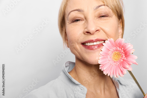 Smiling mature woman holds pink gerbera flower near face, wearing light shirt on white background. Concept of happiness, nature, beauty, and positive lifestyle for seniors.