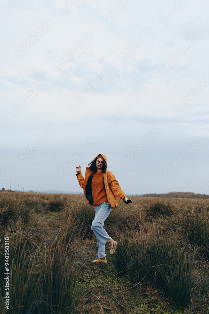 Fototapeta premium Young woman lifestyle walking outdoors in casual autumn clothing with coffee cup in hand. Natural environment with grass and cloudy sky creates relaxed and cozy mood for daily routine.