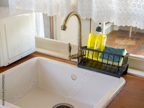 Brass kitchen faucet with white sink and dishwashing supplies on wooden countertop. Clean and organized home kitchen interior with natural light, hygiene, sanitation and domestic lifestyle concept.