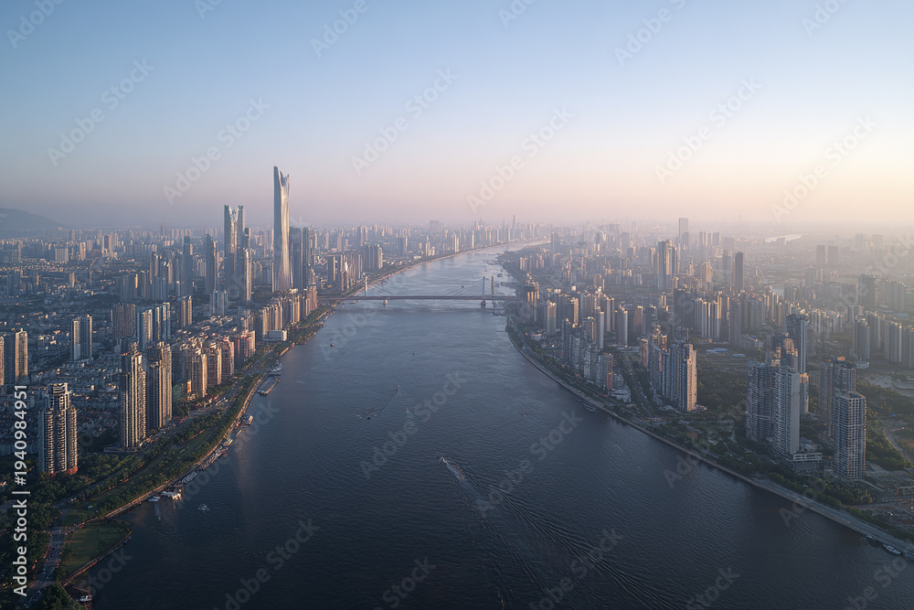 Fototapeta premium Aerial view of modern city skyline with high rise buildings along wide river, bridges crossing water, and hazy sky at sunset creating calm atmosphere