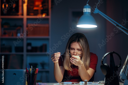 Tableau sur toile Woman with Eyeglasses Struggles to Thread a Needle under Lamp Light