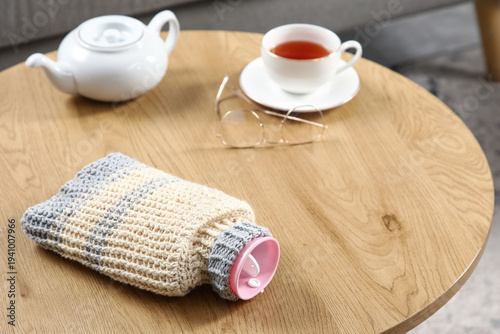 Fotografija Hot water bottle with cup of tea and teapot on coffee table in living room