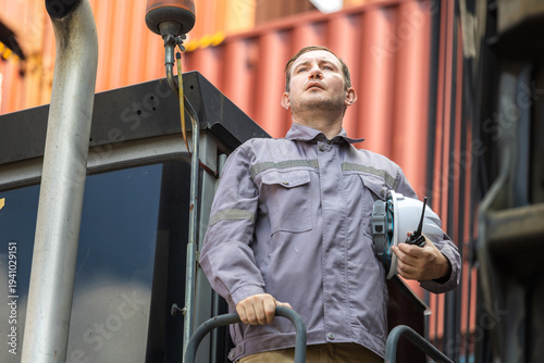 Logistics worker with radio and hardhat at container terminal, professional truck driver or operator standing on vehicle in shipping port